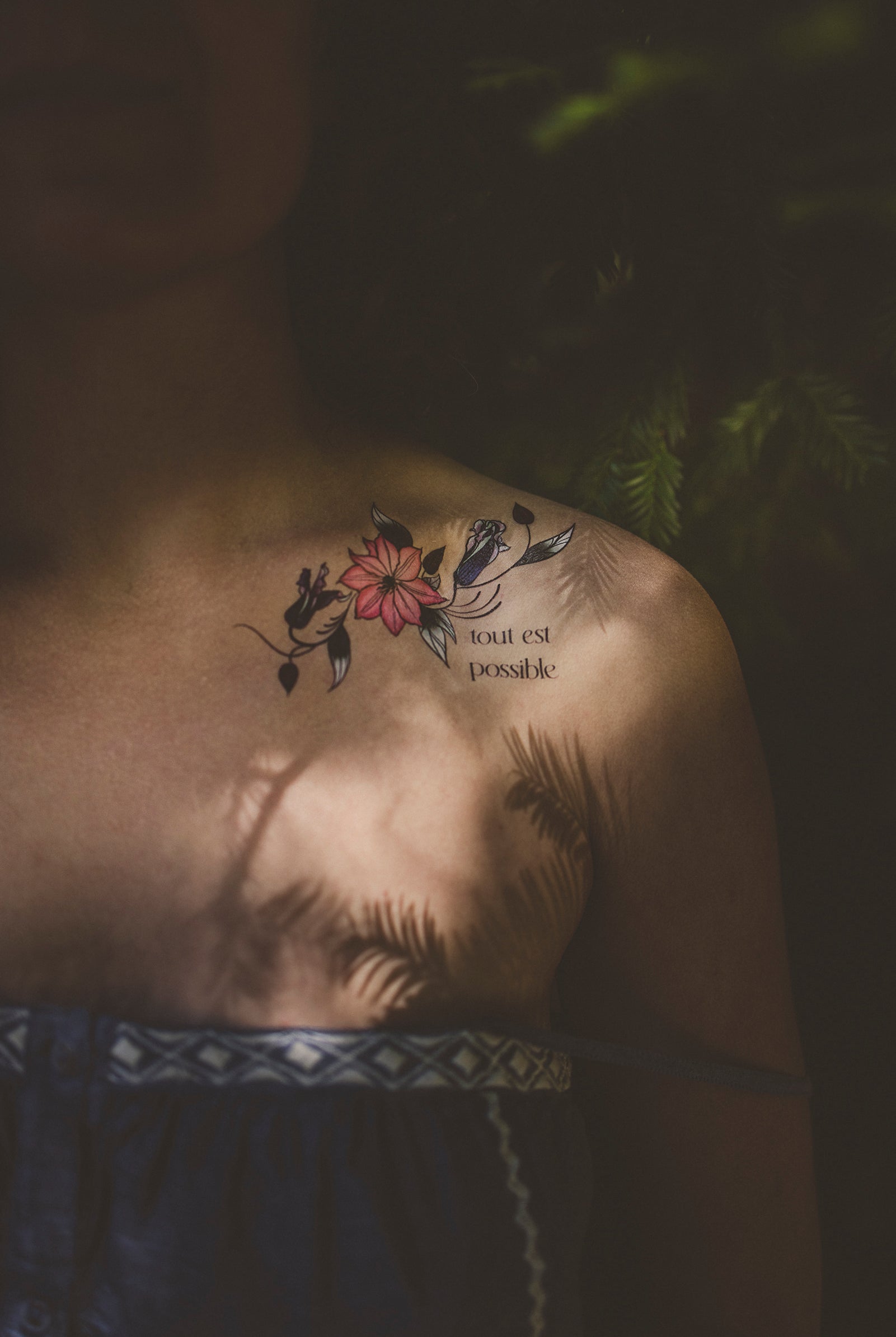 Person wearing a "Tout Est Possible" botanical tattoo on their shoulder as a daily affirmation with sun and shadows.